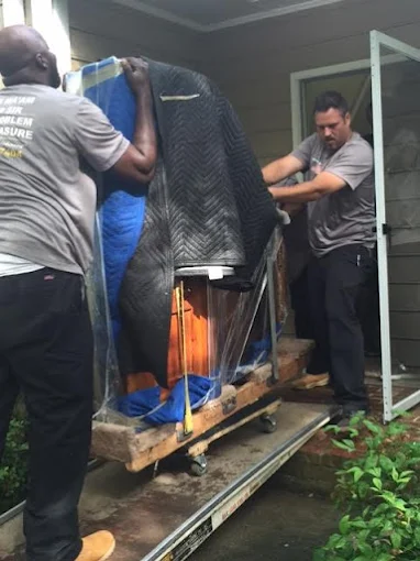 Two professional movers are transporting a large item, wrapped in blue and black moving blankets, up a metal ramp into a building entrance using a wooden dolly.