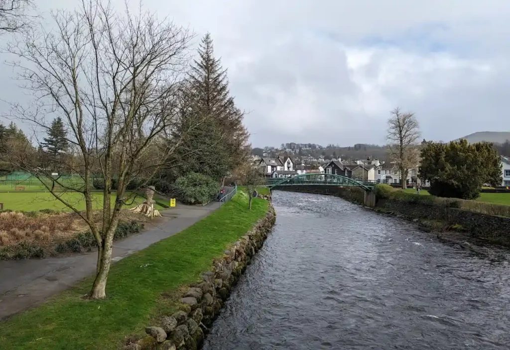 Scenic river with a green bridge and town buildings under a cloudy sky.