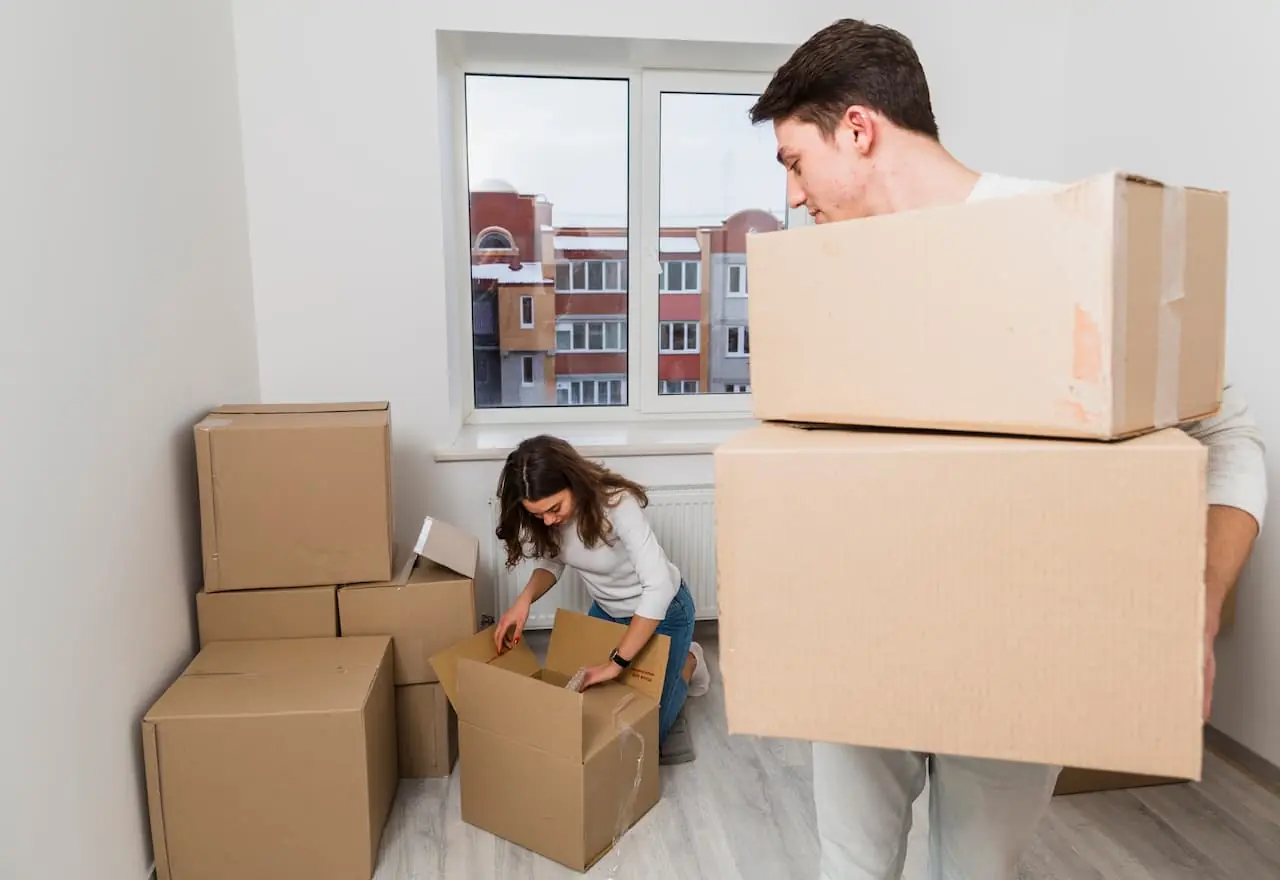 Young couple packing cardboard boxes in a bright, empty apartment room.